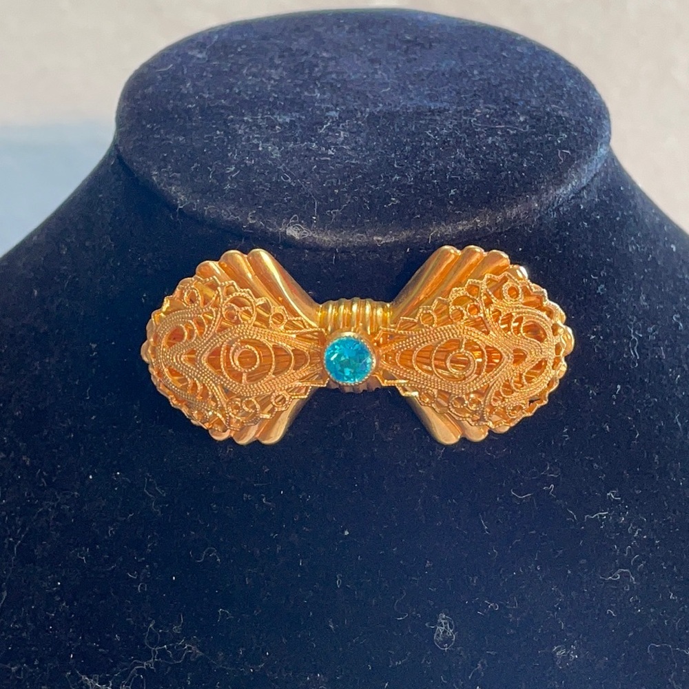 Bow tie brooch gold tone w/ blue glass stone 2.25”. Filigree over shell pattern
